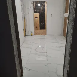 Wnętrze w trakcie prac wykończeniowych: białe płytki podłogowe z marmurkowym wzorem, surowe ściany z widocznymi przewodami, fragment łazienki z czarno-złotymi płytkami i drewnopodobnymi elementami...