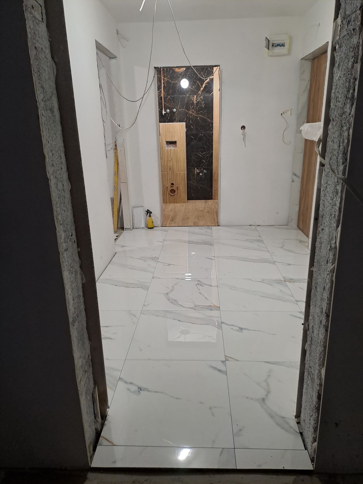 Wnętrze w trakcie prac wykończeniowych: białe płytki podłogowe z marmurkowym wzorem, surowe ściany z widocznymi przewodami, fragment łazienki z czarno-złotymi płytkami i drewnopodobnymi elementami...