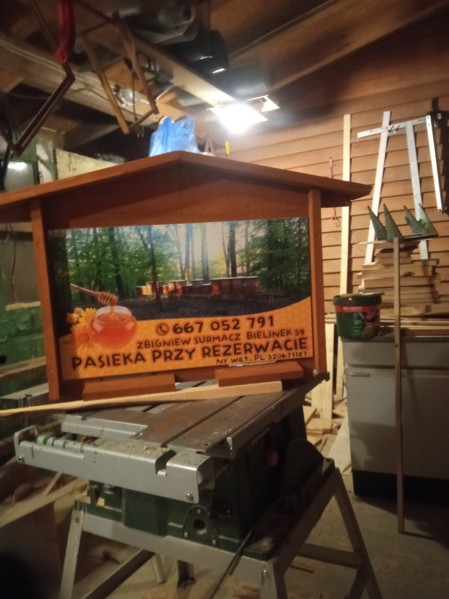 Reklama pasieki z grafiką uli i słoika z nabierakiem, umieszczona na konstrukcji z drewna, stojąca na pile stołowej w warsztacie.