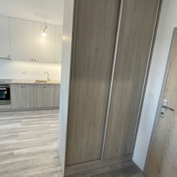 SZWAGRY DESIGN meble z pasją - Szafa wnękowa z przesuwnymi drzwiami w odcieniach szarości, dopasowana do kolorystyki podłogi i kuchni w tle, tworząca spójną aranżację wnętrza.