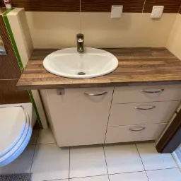 Łazienka z umywalką nablatową na szafce z trzema szufladami i blatem z drewnopodobnym wzorem, obok toaleta, widoczne płytki ścienne w odcieniach beżu i brązu z zielonym akcentem.