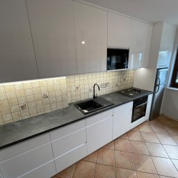 SZWAGRY DESIGN meble z pasją - Nowoczesna kuchnia z białymi, lakierowanymi szafkami, czarnym blatem i zlewozmywakiem, oraz płytkami z motywem owocowym na ścianie.