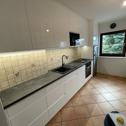 SZWAGRY DESIGN meble z pasją - Nowoczesna kuchnia z białymi, błyszczącymi szafkami wiszącymi i dolnymi, szarym blatem, czarnym zlewem i baterią, oraz beżowymi płytkami ściennymi z motywem owoców; widok na okno z zielonym drzewem...