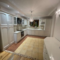 SZWAGRY DESIGN meble z pasją - Elegancka, jasna kuchnia z białymi meblami, ozdobnym żyrandolem i płytkami podłogowymi w odcieniach beżu i brązu. Widoczna zabudowa kuchenna z ażurowymi frontami szafek górnych i blatem z wbudowaną...