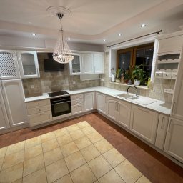 SZWAGRY DESIGN meble z pasją - Jasna kuchnia z białymi szafkami, kryształowym żyrandolem i dwukolorową podłogą z płytek. Widoczne rośliny w oknie.