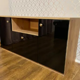 Niska komoda z czarnymi, lakierowanymi frontami i otwartą półką, wykończona okleiną drewnopodobną, z okrągłymi uchwytami w kolorze złota i czerni, na tle ściany w geometryczny wzór.