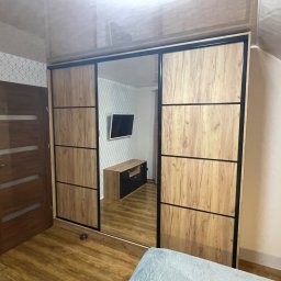 SZWAGRY DESIGN meble z pasją - Przestronna szafa przesuwna z lustrem, jasne drewno z czarnymi akcentami, odbicie pokoju z telewizorem i szafką RTV, widok na podłogę i fragment ściany z tapetą.
