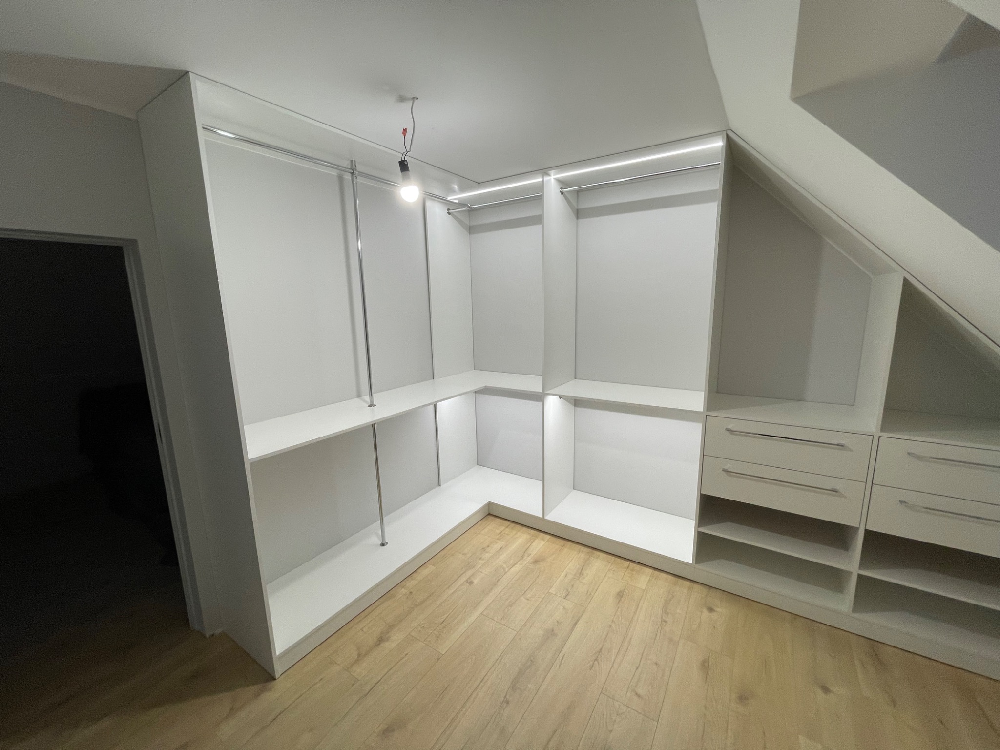Biała garderoba z półkami i drążkami na ubrania, wkomponowana w skos dachu, oświetlona listwą LED, z drewnianą podłogą