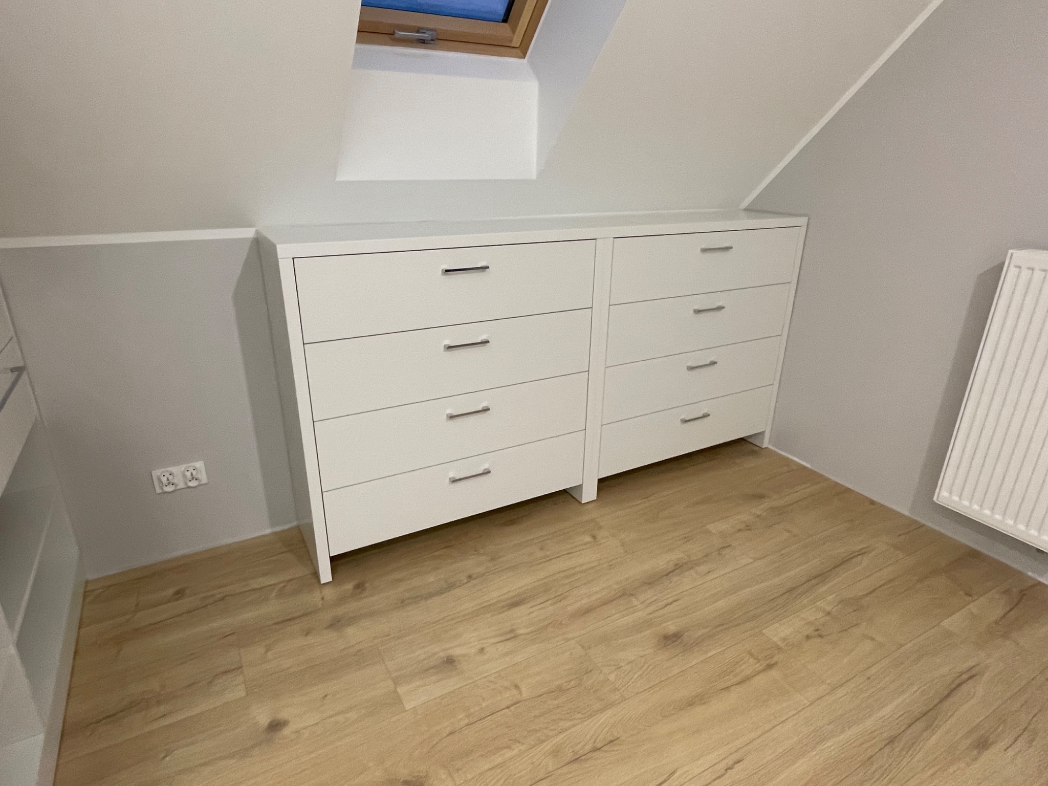 Dwie białe komody z szufladami pod skosem dachu, srebrne uchwyty, panele podłogowe w odcieniach beżu, widoczne gniazdka elektryczne i grzejnik.