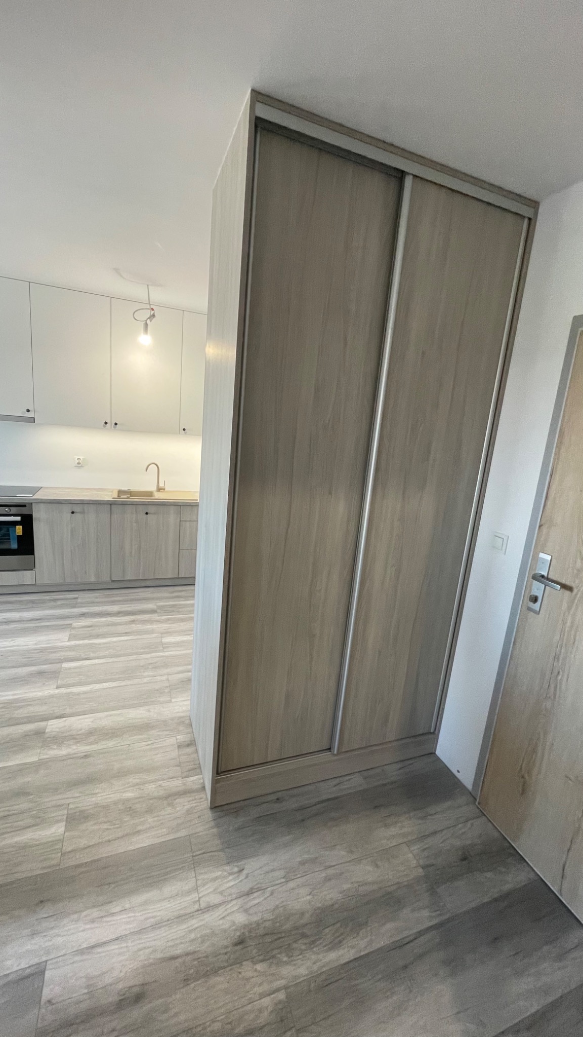 Szafa wnękowa z przesuwnymi drzwiami w odcieniach szarości, dopasowana do kolorystyki podłogi i kuchni w tle, tworząca spójną aranżację wnętrza.
