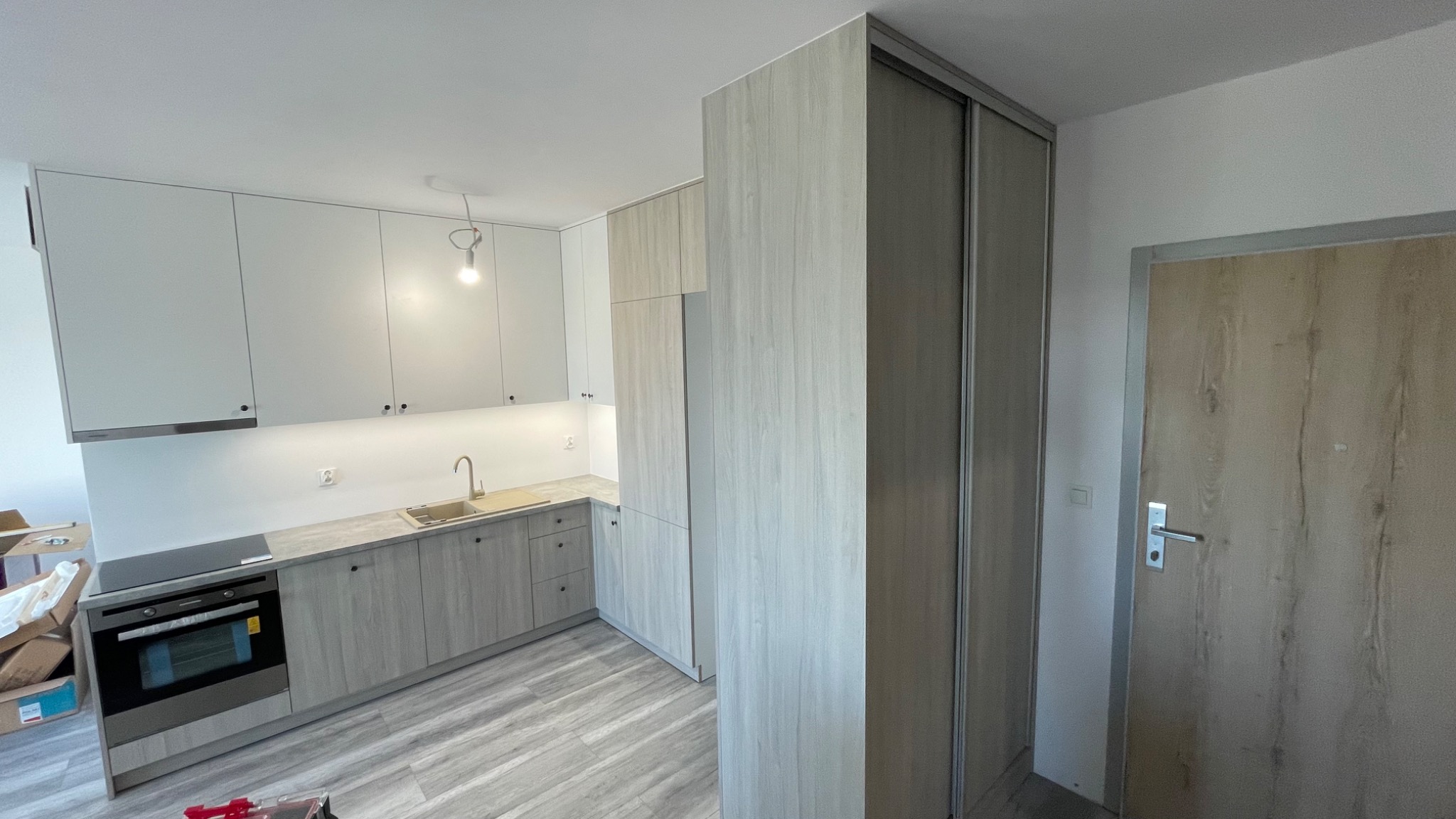 Jasna kuchnia z zabudową sięgającą sufitu, fronty w odcieniach szarości i bieli, widoczny fragment szafy przesuwnej i drzwi wejściowych.