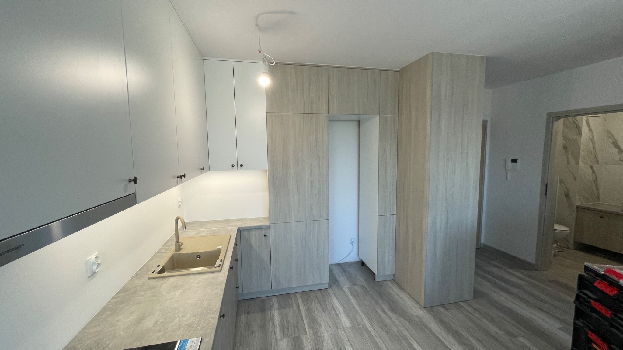 Nowoczesna kuchnia z szarymi i białymi szafkami, beżowym zlewem i szarym blatem, z widocznym wejściem do łazienki w tle.