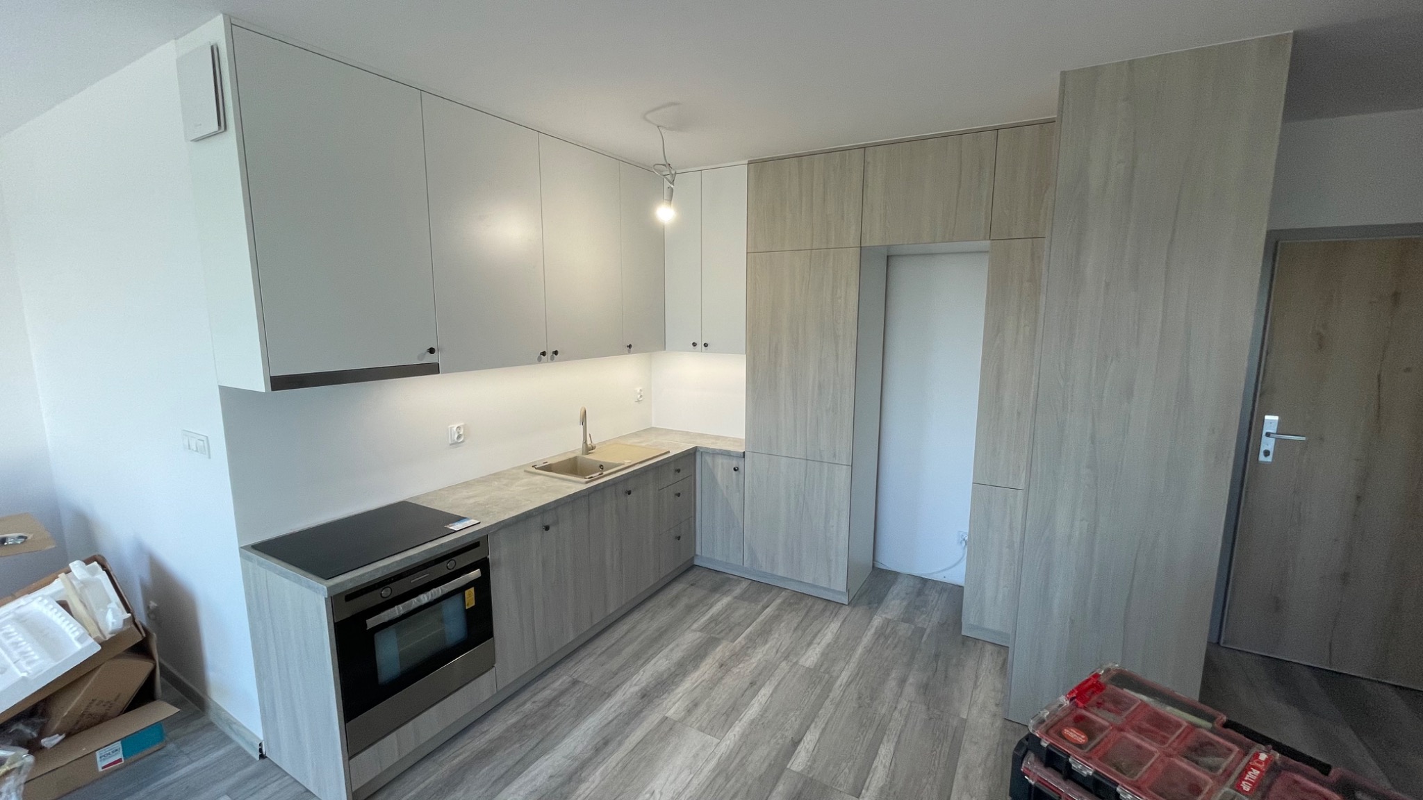 Jasna kuchnia z drewnianymi akcentami, widoczne szafki górne i dolne, płyta indukcyjna, piekarnik, zlew i skrzynka narzędziowa w rogu pomieszczenia.