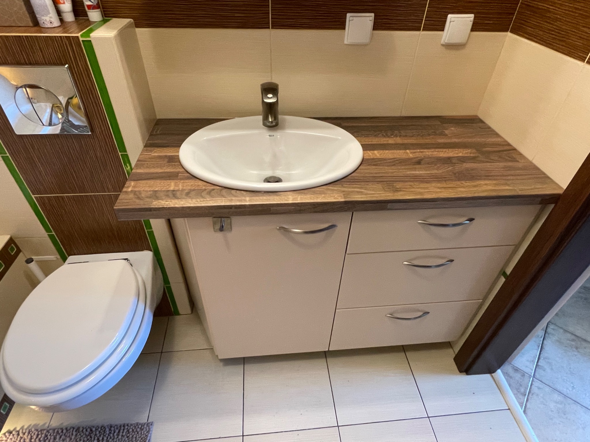 Łazienka z umywalką nablatową na szafce z trzema szufladami i blatem z drewnopodobnym wzorem, obok toaleta, widoczne płytki ścienne w odcieniach beżu i brązu z zielonym akcentem.