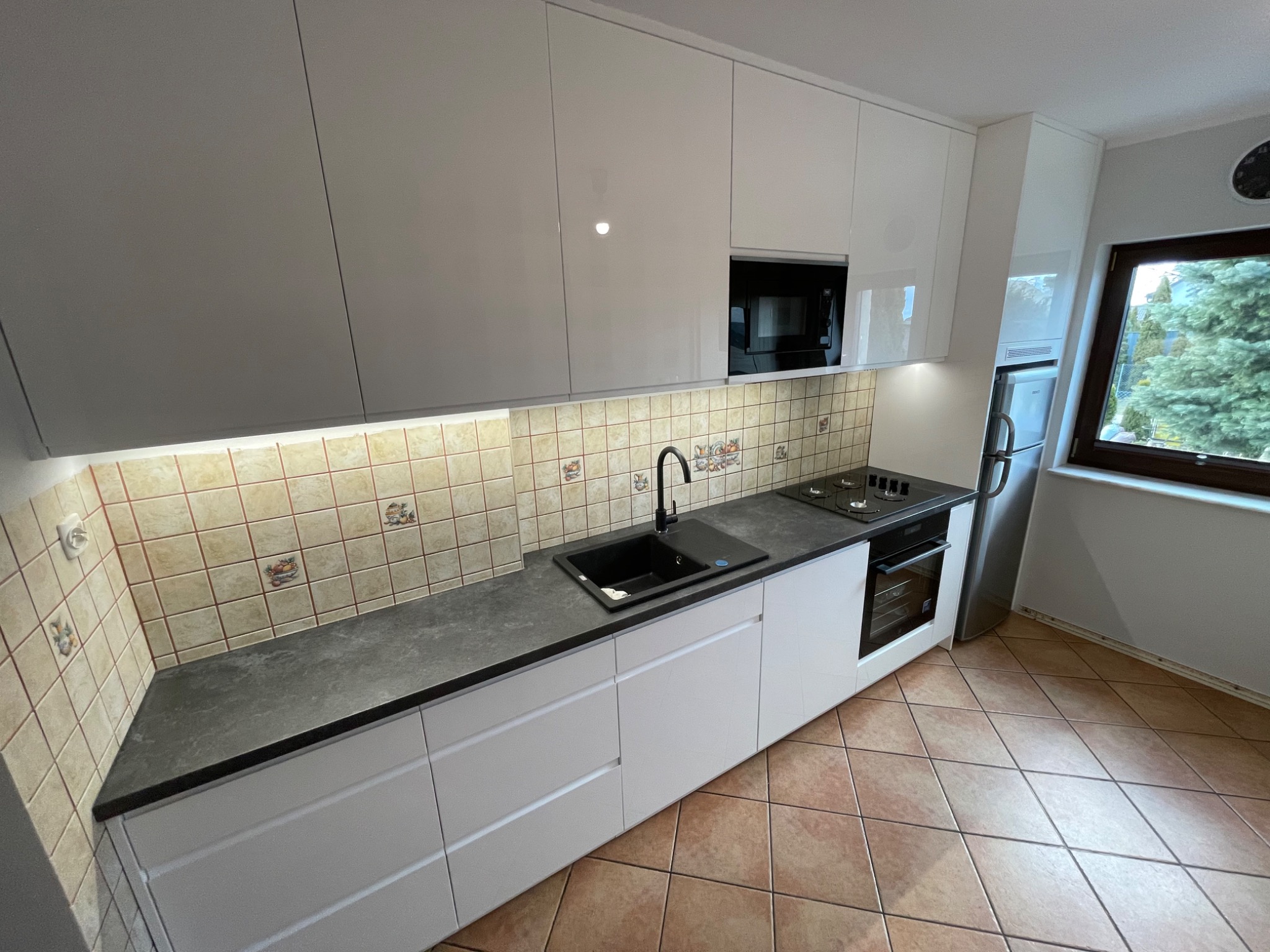 Nowoczesna kuchnia z białymi, lakierowanymi szafkami, czarnym blatem i zlewozmywakiem, oraz płytkami z motywem owocowym na ścianie.