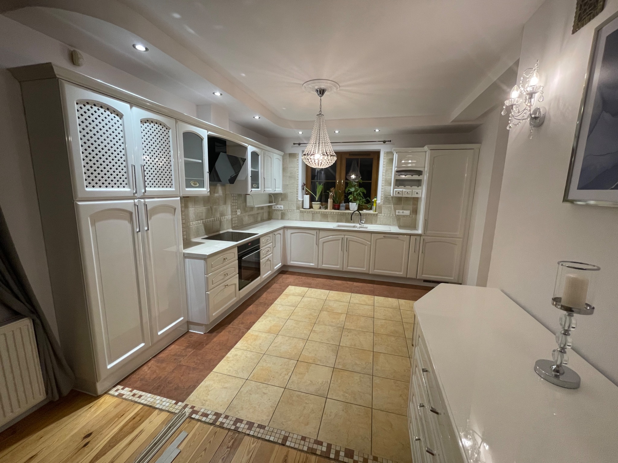 Elegancka, jasna kuchnia z białymi meblami, ozdobnym żyrandolem i płytkami podłogowymi w odcieniach beżu i brązu. Widoczna zabudowa kuchenna z ażurowymi frontami szafek górnych i blatem z wbudowaną...