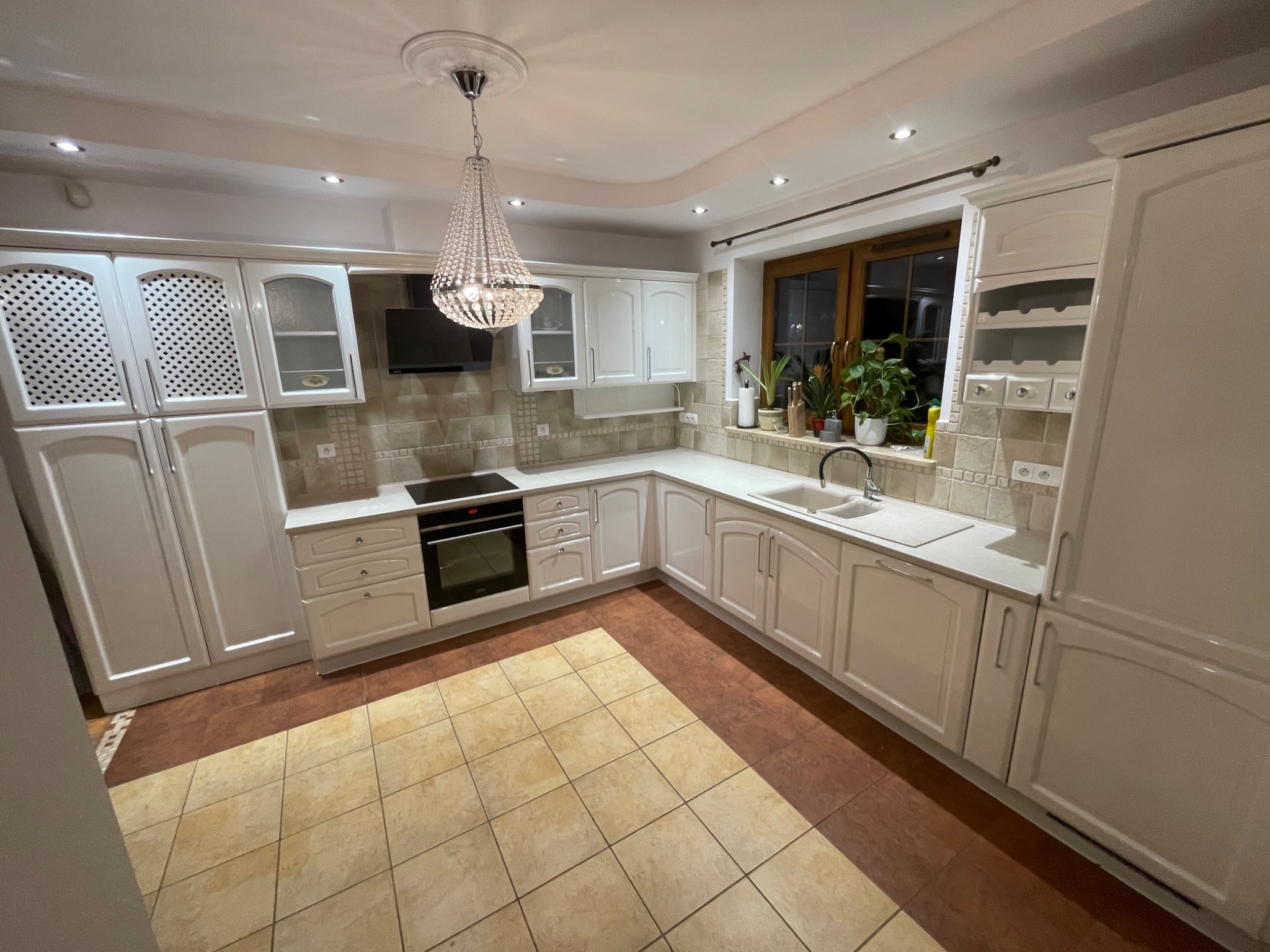 Jasna kuchnia z białymi szafkami, kryształowym żyrandolem i dwukolorową podłogą z płytek. Widoczne rośliny w oknie.