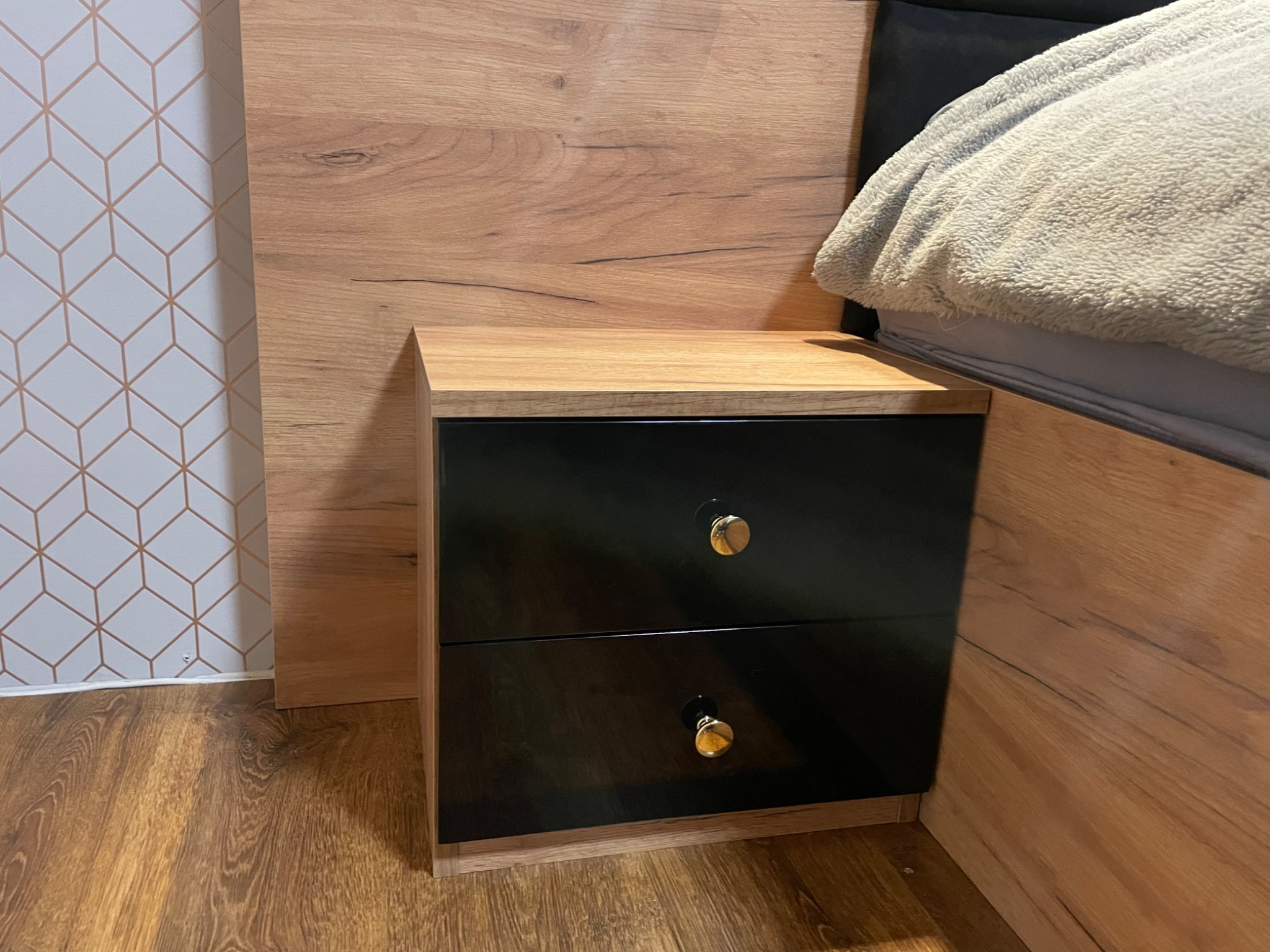 Szafka nocna z dwoma czarnymi, lakierowanymi szufladami i złotymi gałkami, dopasowana do drewnianego wezgłowia łóżka, na tle ściany w geometryczny wzór.