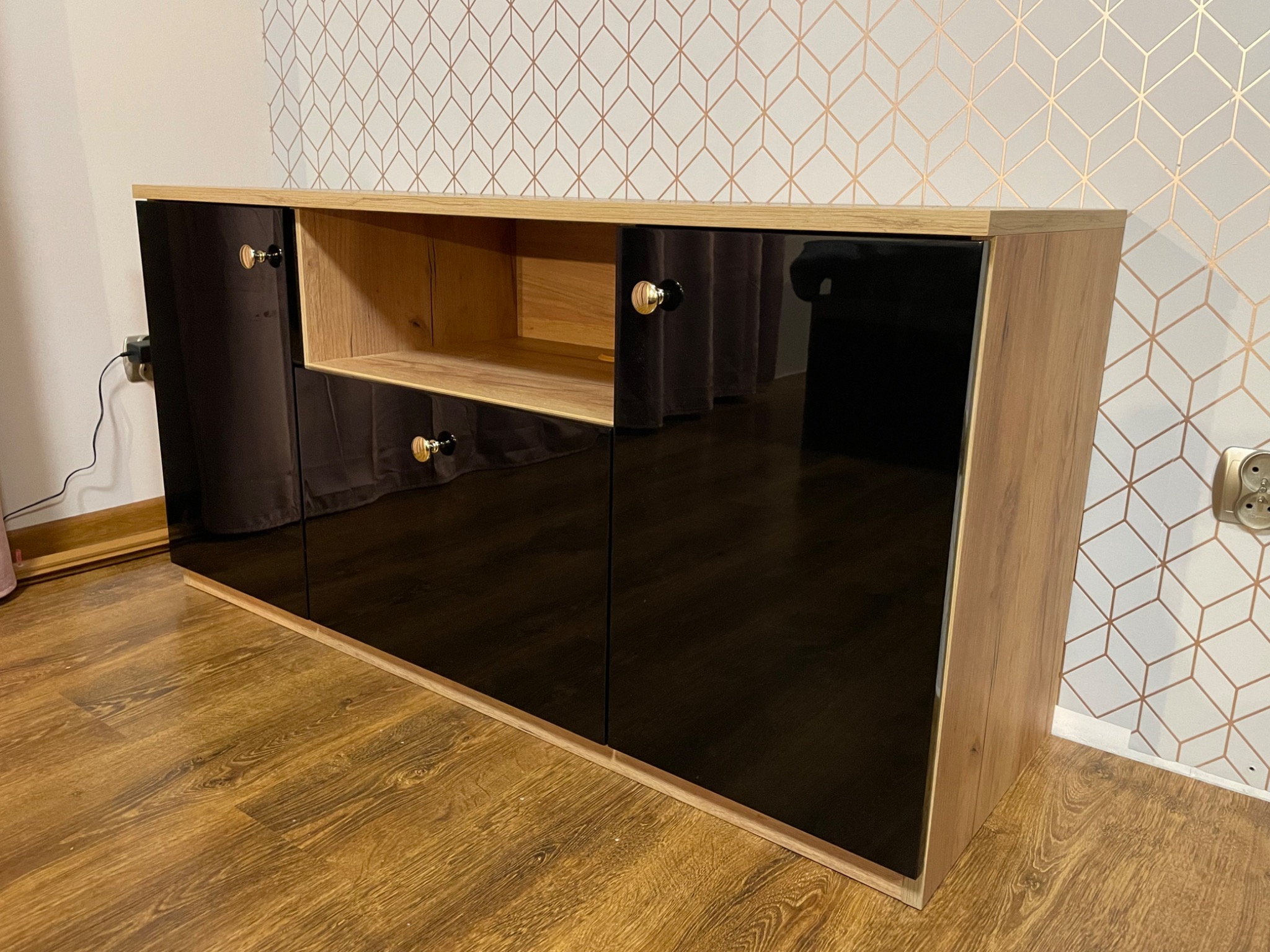 Niska komoda z czarnymi, lakierowanymi frontami i otwartą półką, wykończona okleiną drewnopodobną, z okrągłymi uchwytami w kolorze złota i czerni, na tle ściany w geometryczny wzór.