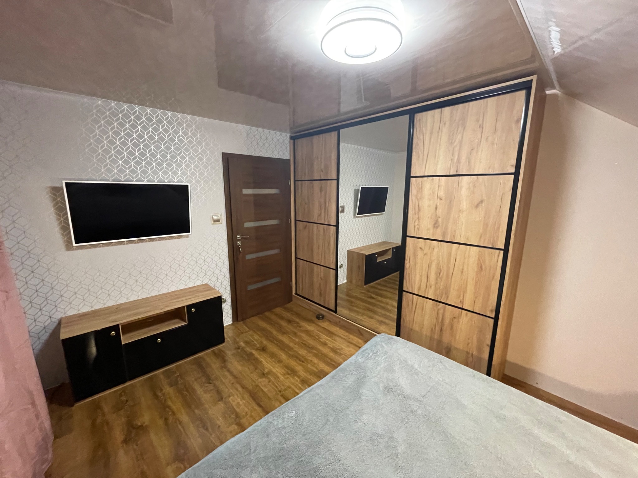 Szafa wnękowa z przesuwnymi drzwiami z lustrem, drewnopodobne fronty z czarnymi akcentami, dopasowana szafka RTV w kolorze czarnym z drewnianymi wstawkami, błyszczący sufit i wzorzysta tapeta...