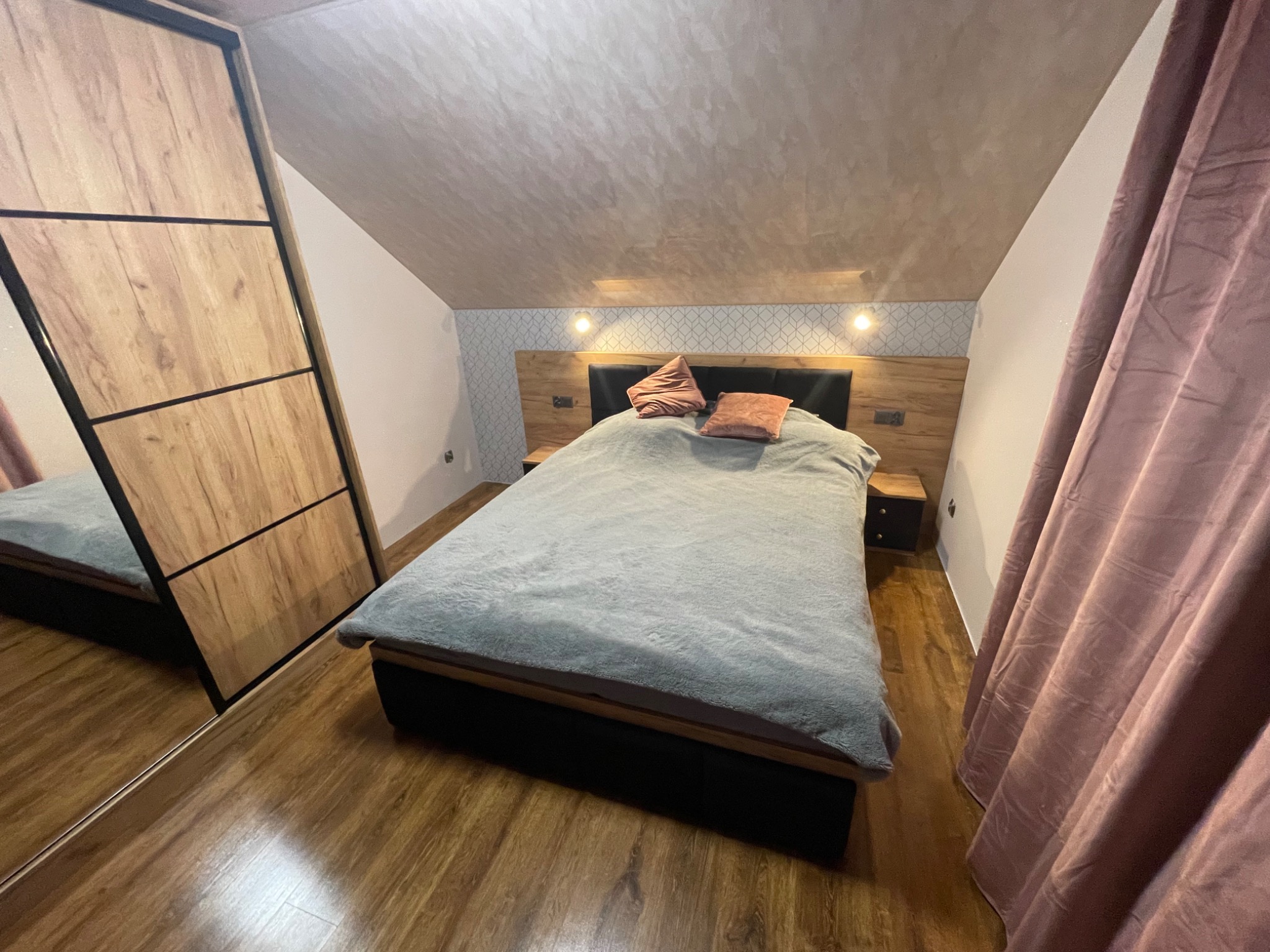 Sypialnia na poddaszu z łóżkiem, szafkami nocnymi i drewnianą szafą przesuwną z lustrem, widok na skośny sufit i zasłony w kolorze pudrowego różu.