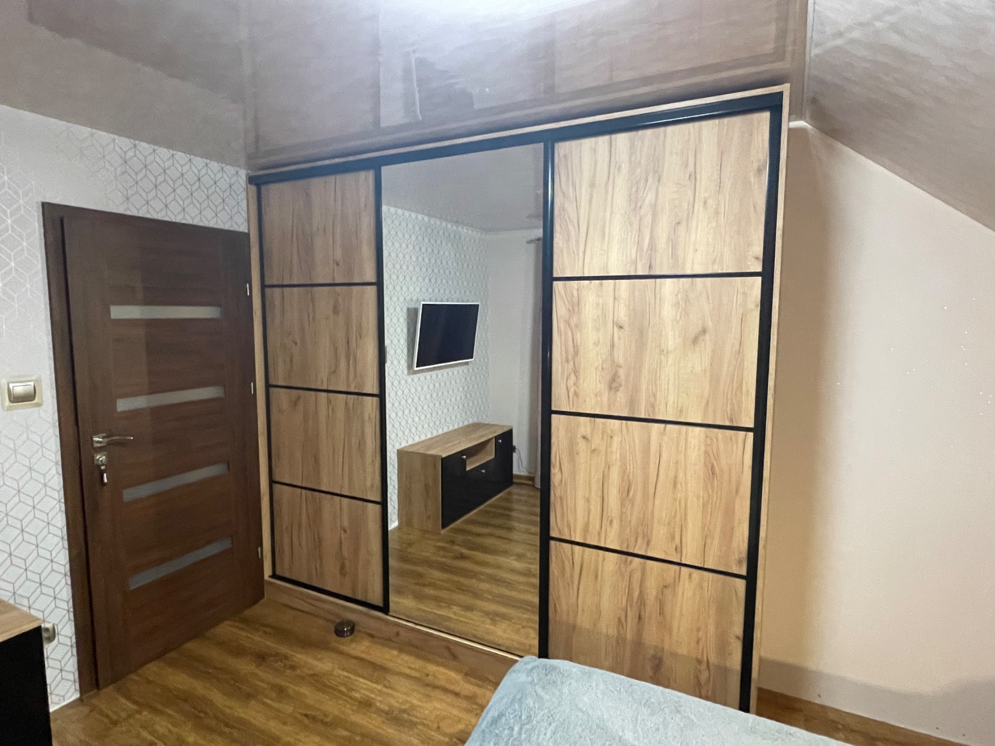 Przestronna szafa przesuwna z lustrem, jasne drewno z czarnymi akcentami, odbicie pokoju z telewizorem i szafką RTV, widok na podłogę i fragment ściany z tapetą.