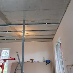 Montaż metalowego stelażu pod płytę gipsowo-kartonową na suficie, widoczne profile i konstrukcja nośna, w tle okno i narzędzia na półce.