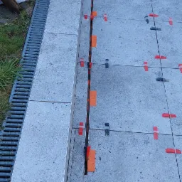 Układanie szarych płytek na zewnątrz z użyciem pomarańczowych i czarnych klipsów poziomujących, obok odpływu liniowego.