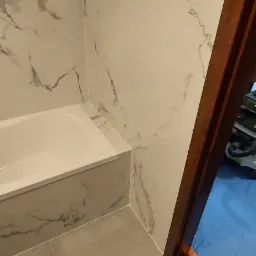 Wykończona łazienka z wanną obudowaną płytkami imitującymi marmur, szare płytki podłogowe, widok na drewnianą futrynę drzwi i niebieską folię ochronną na podłodze w sąsiednim pomieszczeniu.