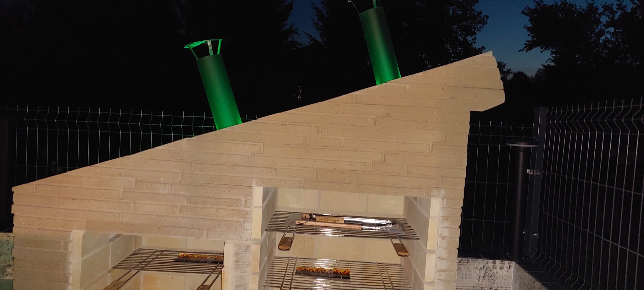 Murowany grill z cegły z zielonymi kominami, oświetlony w nocy, z rusztem i akcesoriami do grillowania w środku.