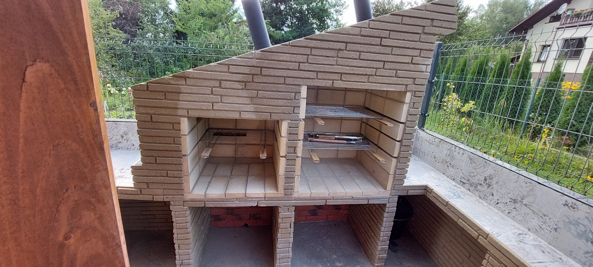 Murowany grill z cegły klinkierowej z dwoma komorami, z rusztem w jednej z nich, na zewnątrz, widok z boku, z elementami drewnianymi po lewej stronie kadru oraz metalową siatką ogrodzeniową w tle.