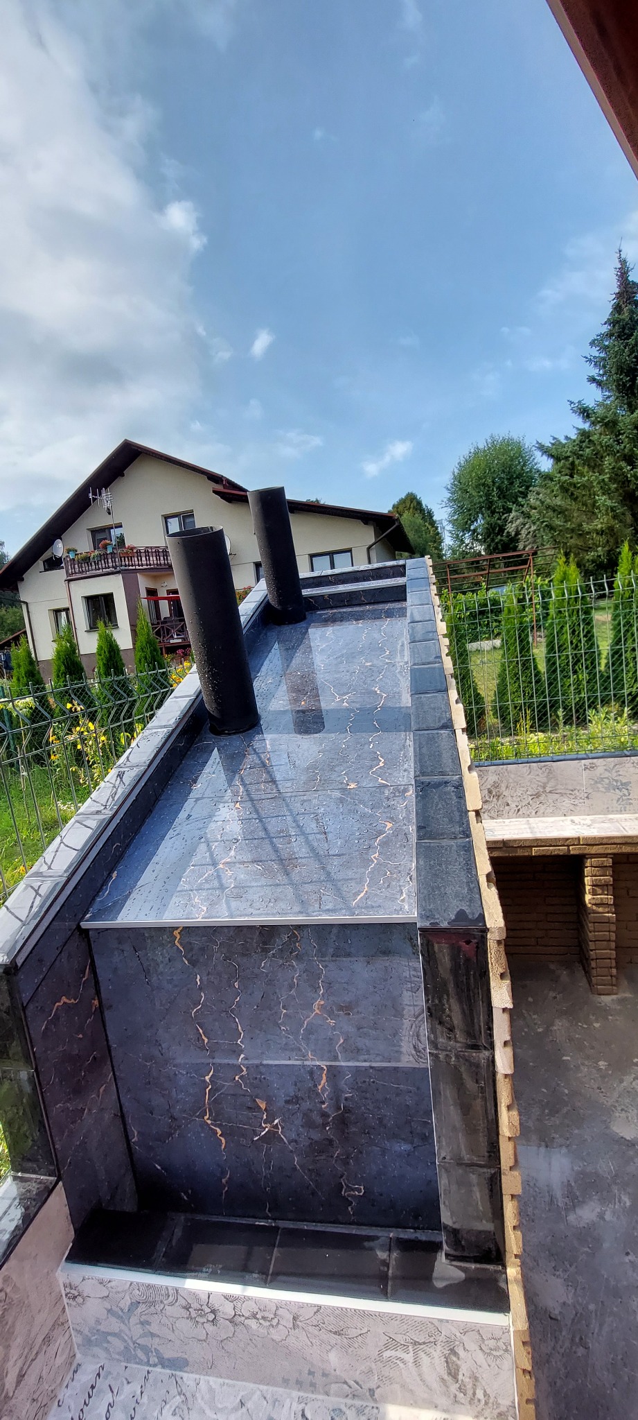 Wykończony grill ogrodowy z czarnymi płytkami imitującymi marmur, z dwoma kominami, na tle domu i zieleni.