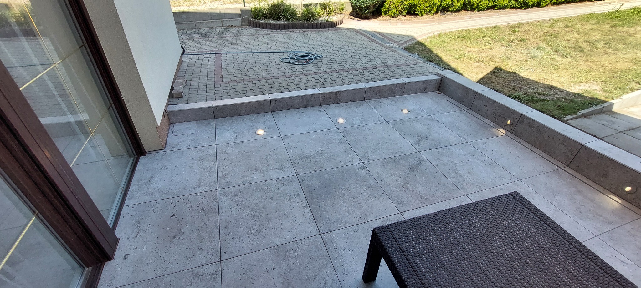 Wykończony taras z szarych płytek, wbudowane oświetlenie punktowe w posadzce i ścianie, widok na ogród i kostkę brukową, fragment mebla ogrodowego w rogu.