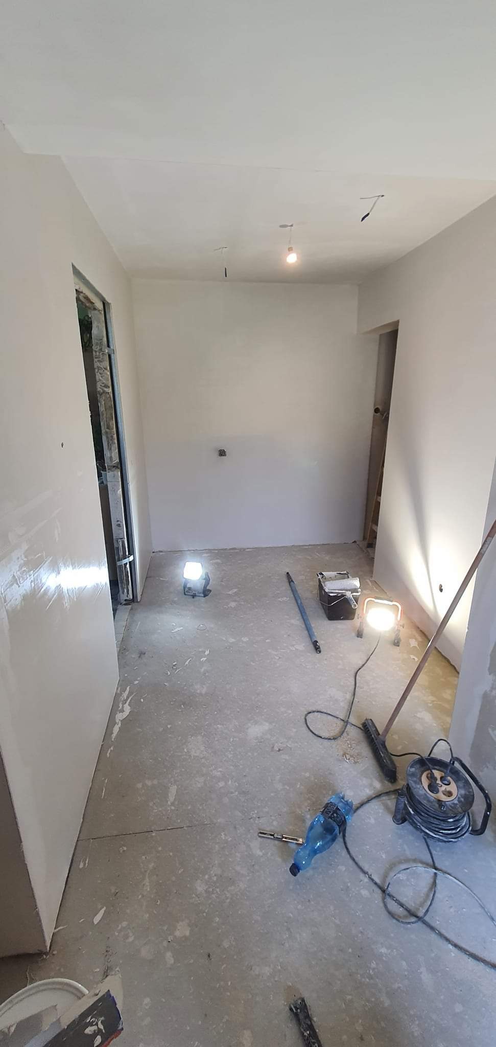 Wnętrze w trakcie prac wykończeniowych: ściany pokryte świeżo nałożoną gładzią, widoczne halogeny budowlane oświetlające pomieszczenie, narzędzia malarskie i butelka wody na podłodze.