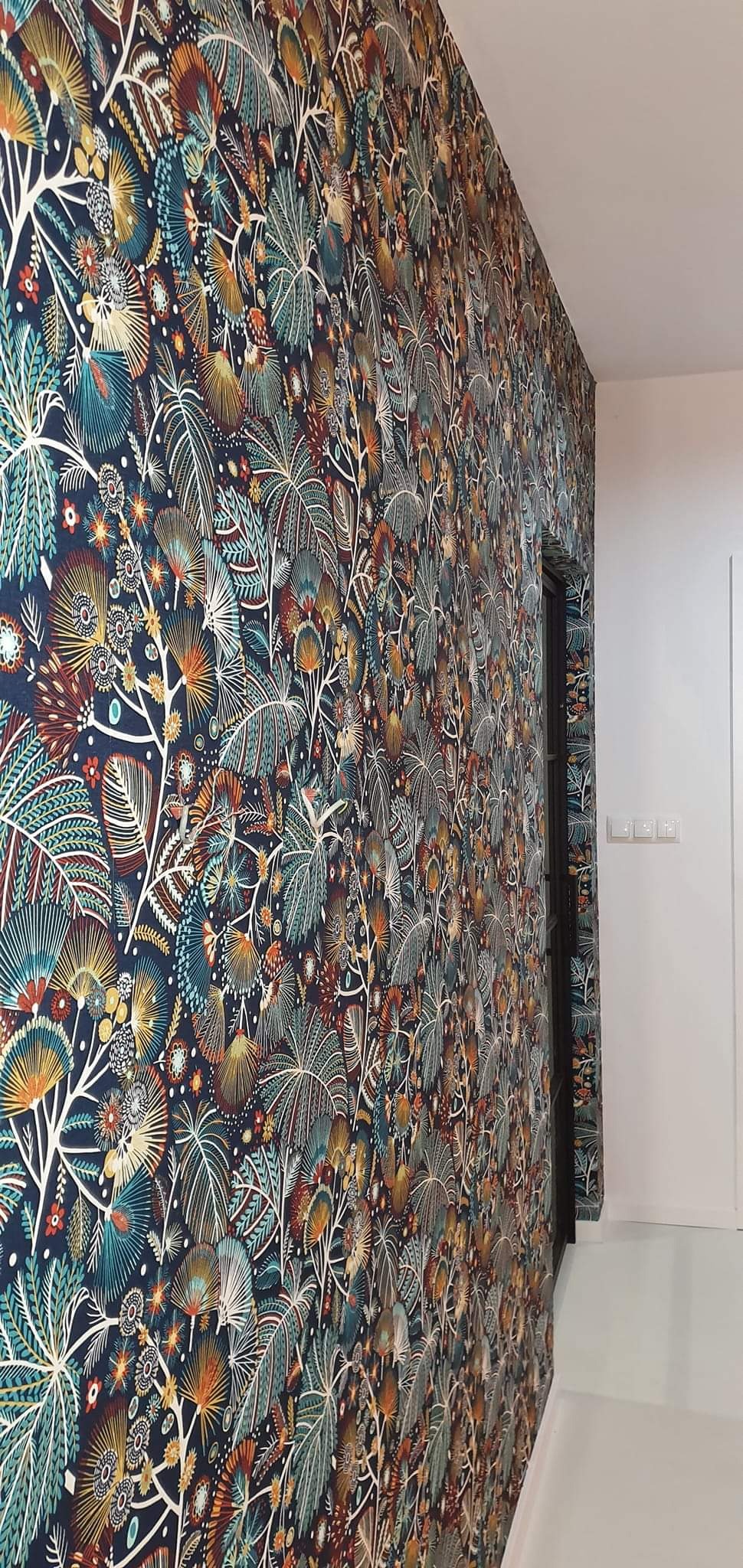 Ściana pokryta wzorzystą tapetą w stylu tropikalnym, z motywami liści i kwiatów w odcieniach granatu, turkusu, pomarańczy i brązu, sąsiadująca z białą ścianą i czarnymi drzwiami.
