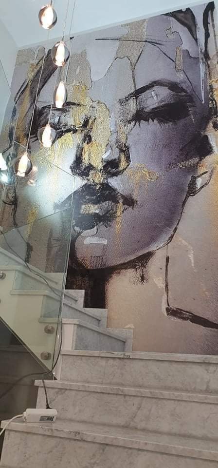 Abstrakcyjny portret kobiety w odcieniach szarości i złota na ścianie przy marmurowych schodach ze szklaną balustradą i wiszącymi lampami.