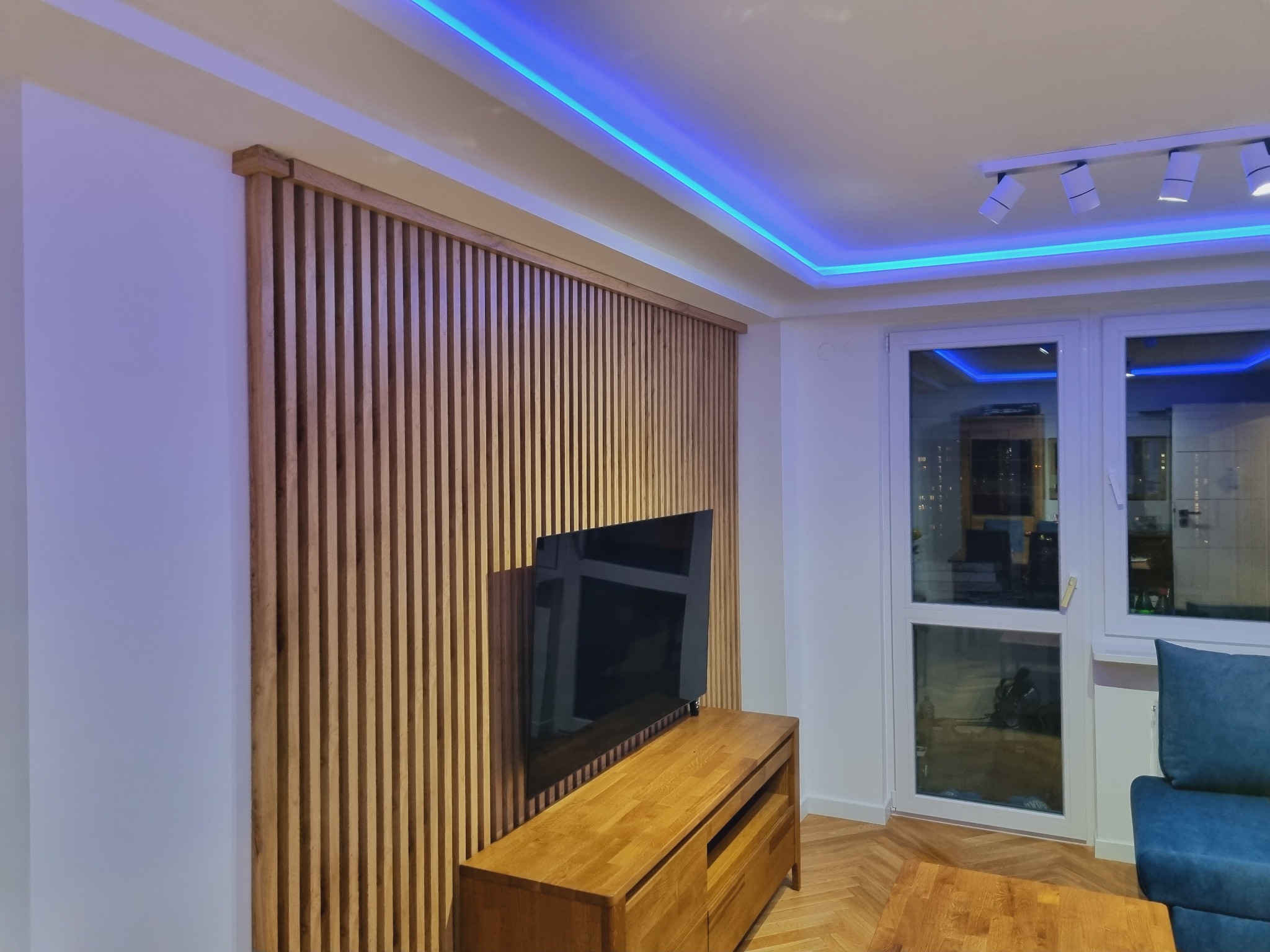 Drewniana lamelowa ściana za telewizorem, podświetlona niebieskim oświetleniem LED, z drewnianą szafką RTV i parkietem w jodełkę.