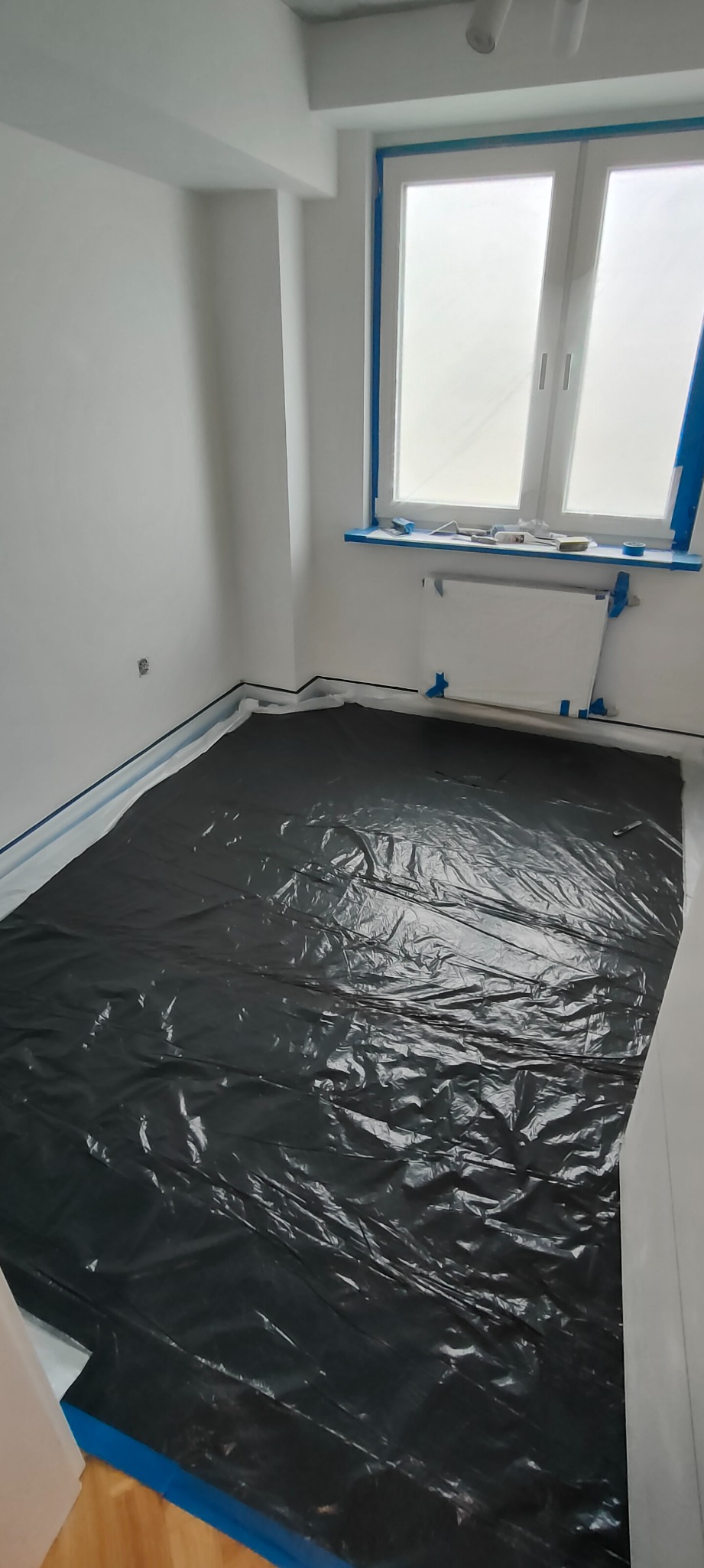 Pokój przygotowany do malowania: podłoga zabezpieczona czarną folią, okna i listwy przypodłogowe oklejone niebieską taśmą malarską, ściany białe, grzejnik osłonięty folią ochronną.