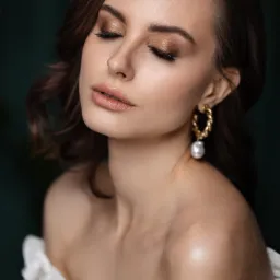 Portret kobiety z zamkniętymi oczami, delikatny połyskujący cień do powiek w odcieniach złota, błyszczące usta, złote kolczyki z perłą i biała bluzka z falbankami na tle ciemnej zieleni.