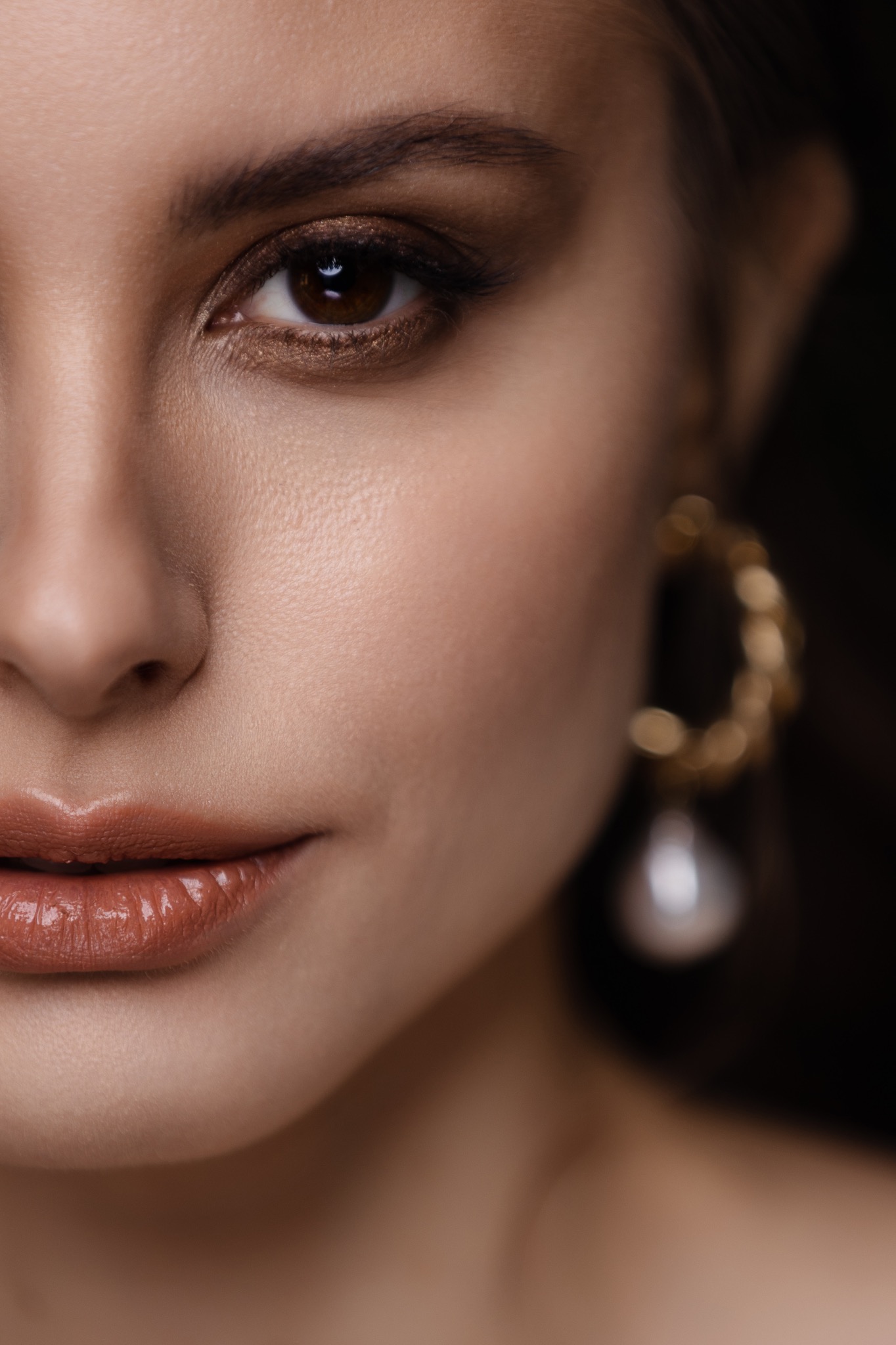 Detal portretu kobiety z błyszczącym, brązowym cieniem na powiekach, podkreślonym okiem i błyszczykiem na ustach, z widocznym złotym kolczykiem z perłą.