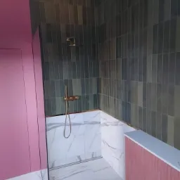 Nowoczesna łazienka z pionowo ułożonymi, grafitowymi płytkami w strefie prysznica, kontrastującymi z marmurowymi płytkami na podłodze i dolnej części ścian oraz różowymi elementami zabudowy.