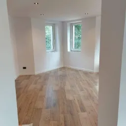 Jasne wnętrze po remoncie z drewnopodobną podłogą, białymi ścianami i dwoma oknami w narożniku, widoczne punktowe oświetlenie sufitowe.