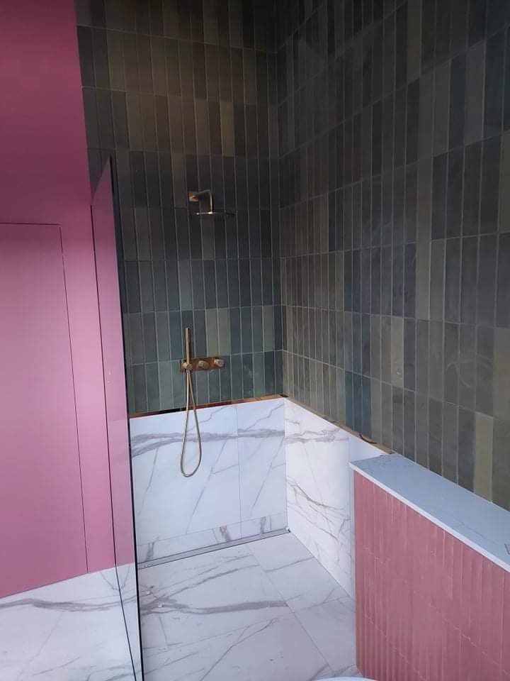 Nowoczesna łazienka z pionowo ułożonymi, grafitowymi płytkami w strefie prysznica, kontrastującymi z marmurowymi płytkami na podłodze i dolnej części ścian oraz różowymi elementami zabudowy.