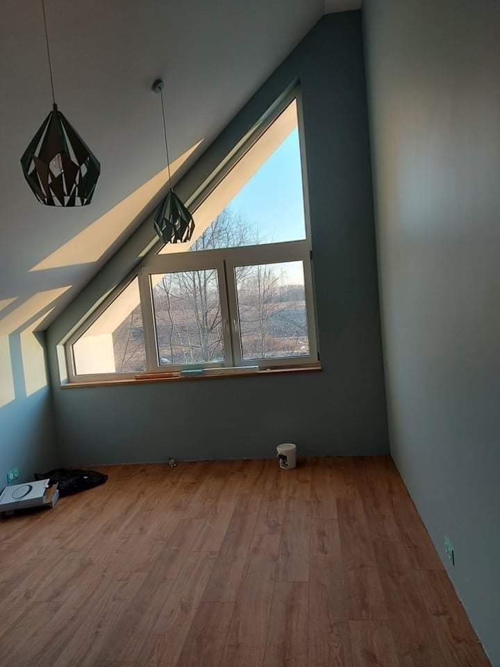 Pomieszczenie na poddaszu po malowaniu ścian na szaro, z drewnianą podłogą, oknem w kształcie trójkąta i dwoma nowoczesnymi lampami wiszącymi.