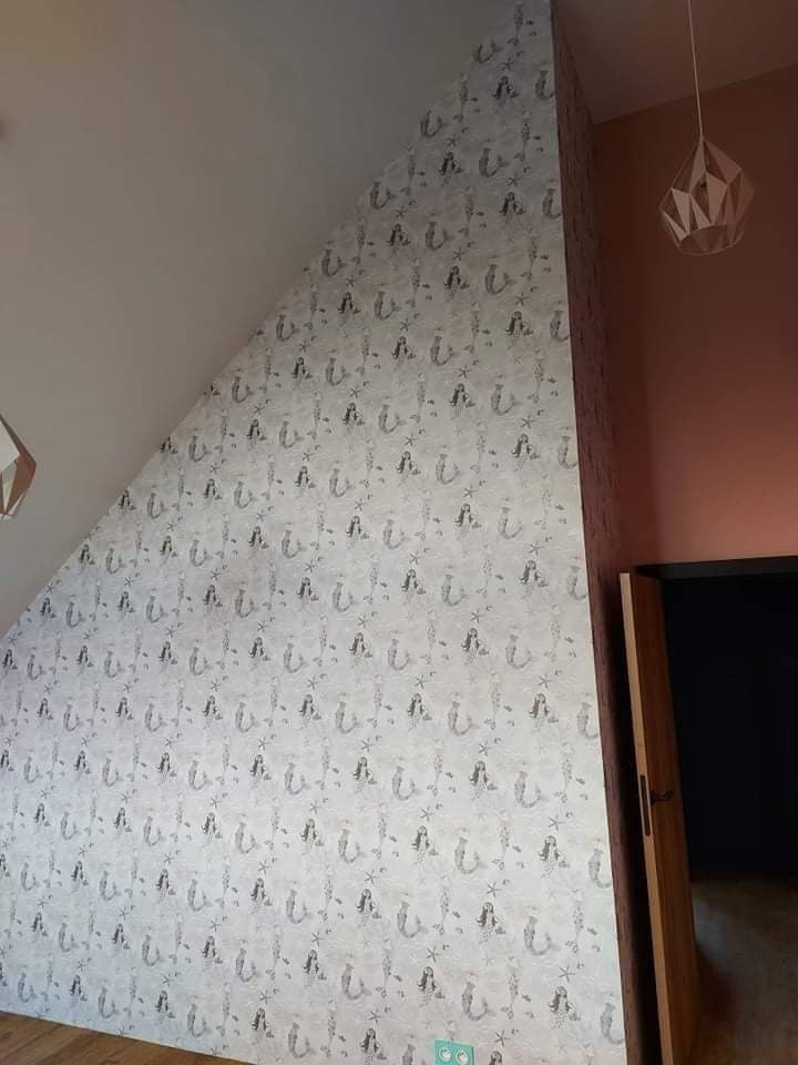 Ściana na poddaszu pokryta tapetą w motywie syrenek, z widoczną lampą wiszącą i fragmentem drzwi.