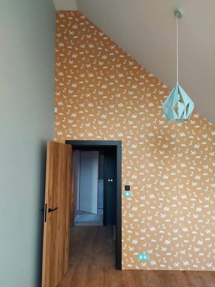 Pokój dziecięcy po remoncie: ściana w kolorze musztardowym z białym wzorem zwierząt, drewniane drzwi z czarną klamką, turkusowa lampa wisząca, szara ściana obok drzwi.