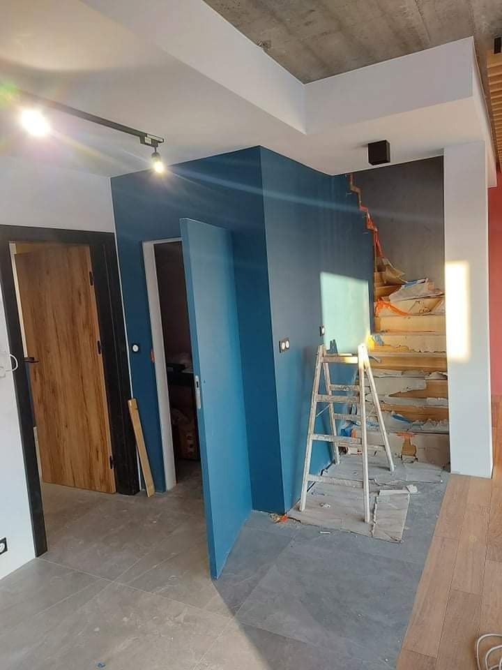 Wnętrze w trakcie remontu: ściany pomalowane na niebiesko, częściowo wykończone schody, drabina oparta o ścianę, widoczne prace wykończeniowe.