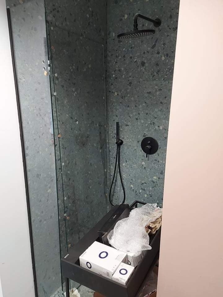 Nowoczesna kabina prysznicowa z zielonymi płytkami imitującymi lastryko, czarną armaturą i szklanymi drzwiami, w trakcie montażu widoczne kartony i folia ochronna.