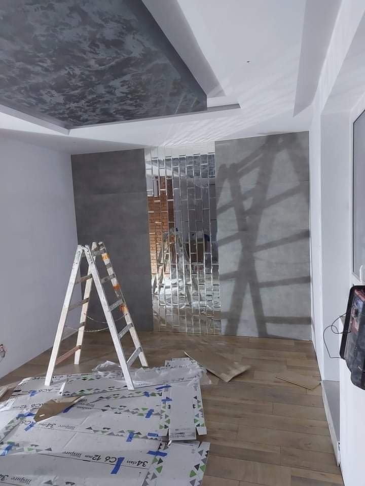 Wnętrze w trakcie prac wykończeniowych: ściana z szarych paneli i lustrzanych płytek ułożonych w cegiełkę, sufit z dekoracyjną szarą fakturą, drewnopodobna podłoga, biała drabina oparta o ścianę...