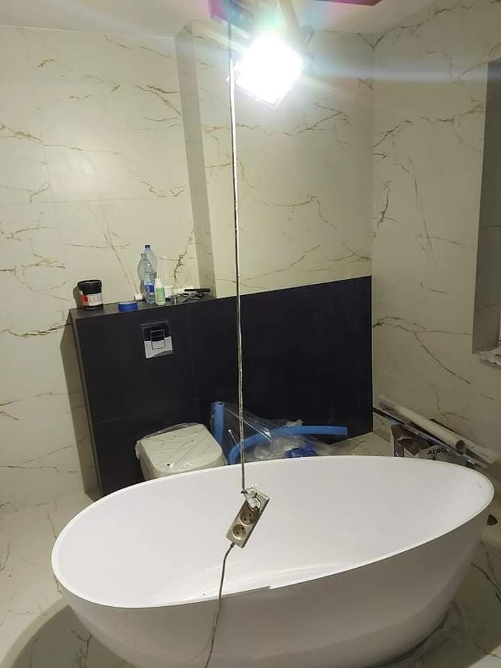 Biała wanna wolnostojąca w trakcie montażu w łazience z marmurowymi płytkami, oświetlona lampą roboczą zawieszoną na kablu z gniazdkiem, widoczny czarny element zabudowy pod umywalkę i akcesoria...